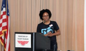 Alicia M Morgan on stage in the auditorium of Frontiers of Flight Museum in Dallas. There is a black podium with red logo and words in black.