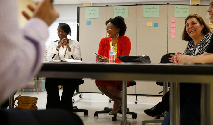 Three women sitting at a table with presenter in front of the room.  