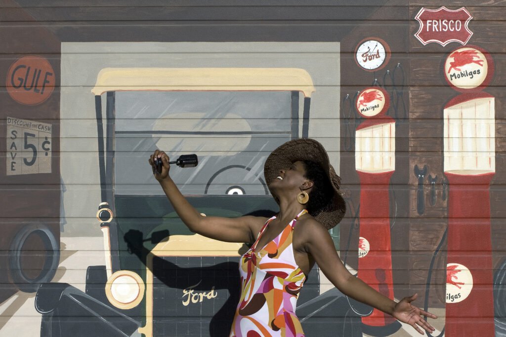 Black Woman with hat holding a microphone with a background mural of cars. 