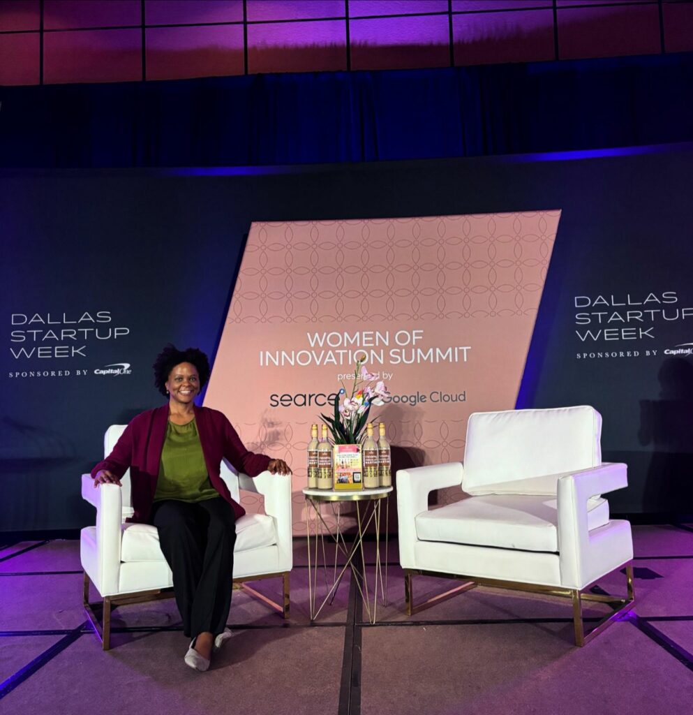 Black Woman sitting in white chairs on a stage with background for Dallas Startup Week Women of Innovation Summit.