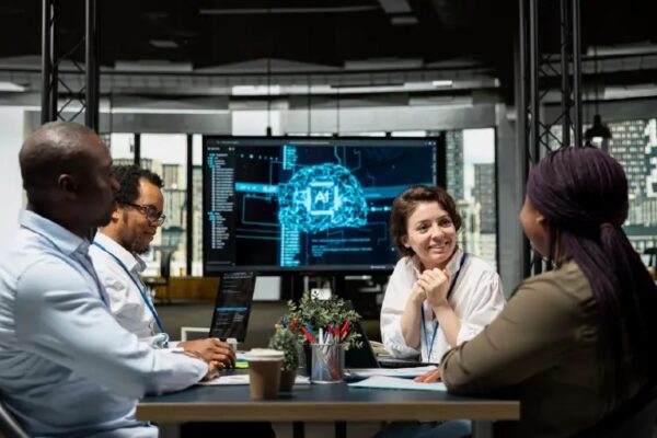 A diverse group of four professionals collaborates around a table in a modern office. They are smiling and engaged in conversation. In the background, a large digital screen displays a glowing blue graphic of a human brain with "AI" in the center, representing the Human-in-the-Loop approach to technology transformation. Laptops and coffee cups are on the table, highlighting a blend of human connection and strategic AI integration.